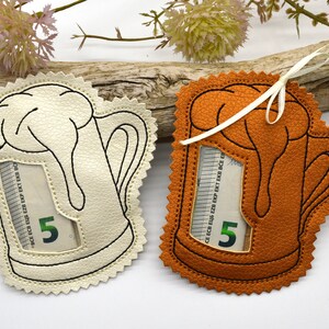 May include: Two felt beer mug shaped money holders with a window to insert a bill. One is white with black stitching and the other is orange with black stitching. Both have a five euro bill inserted.