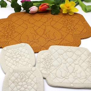 May include: A set of Easter-themed quilted placemats and coasters. The large, mustard-colored placemat features two rabbits and floral designs. The smaller coasters are off-white with floral and rabbit motifs. The set is decorated with a diamond pattern.