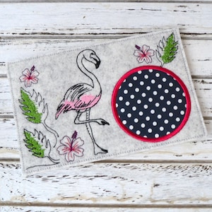 Könnte beinhalten: Ein grauer Filzuntersetzer mit einem rosa Flamingo, grünen Blättern und rosa Hibiskusblüten. Der Untersetzer hat einen schwarzen und weißen Kreis mit Punkten und einem roten Rand.