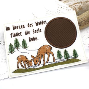 May include: Embroidered patch featuring two brown deer in a snowy landscape with green pine trees. The patch has a brown circular area and text that reads "Im Herzen des Waldes findet die Seele Ruhe."