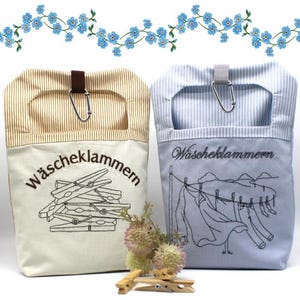 Può includere: Due sacchetti in tessuto per mollette, uno beige con la parte superiore a righe marroni e uno azzurro con la parte superiore a righe blu. Entrambi presentano un moschettone in metallo, la scritta "Wäscheklammern" e disegni a tratto nero.