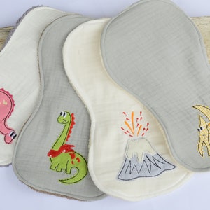 May include: Four white muslin burp cloths with different embroidered dinosaur designs. The designs include a pink dinosaur, a green dinosaur, a volcano, and a pterodactyl.