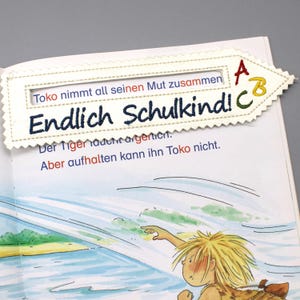 Könnte beinhalten: Ein weißes Stofflesezeichen mit den Worten "Toko nimmt all seinen Mut zusammen" und "Endlich Schulkind!" in dunkelblauer Stickerei. Das Lesezeichen hat eine dekorative Naht und ABC-Buchstaben. Das Bild zeigt eine Seite aus einem Kinderbuch.