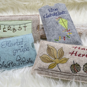 Könnte beinhalten: Vier Filz-Handyhüllen mit herbstlichen Stickereien. Die Hüllen zeigen Motive wie einen Pilz-Patch, ein Blatt, einen Drachen und eine Kastanie. Der Text auf den Hüllen lautet "Herbst", "Liebe Kleinigkeit", "Hallo Herbst!" und "Supe Herbst-grüße".