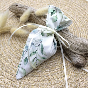 Puede incluir: Una bolsa de tela blanca con un estampado de hojas botánicas en tonos verdes y azules. La bolsa está atada con una cinta blanca. El fondo incluye madera flotante y plantas secas, sugiriendo un tema natural.