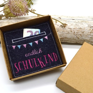 Puede incluir: Una pequeña caja marrón con un inserto de fieltro. El fieltro es azul oscuro con un bolsillo y un mensaje bordado en rosa que dice "endlich SCHULKIND". Hay pequeñas banderas azules y rosas cosidas sobre el bolsillo. Un billete de veinte euros es visible en el bolsillo.