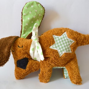 Puede incluir: Un juguete de peluche de perro marrón con una estrella verde y blanca en la espalda. El perro tiene un lazo verde y blanco de lunares atado alrededor del cuello y una oreja con estampado de estrellas verdes y blancas.