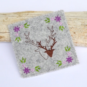 Puede incluir: Un posavasos cuadrado de fieltro gris con una cabeza de ciervo bordada en hilo marrón. El posavasos está decorado con un borde de flores moradas y rosas y hojas verdes. Los bordes tienen un acabado decorativo festoneado.