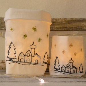 Könnte beinhalten: Zwei weiße Stoff-Kerzenhalter mit gestickten Winterdorfszenen. Der größere Halter zeigt eine Kirche, Bäume und Sterne. Der kleinere Halter zeigt eine ähnliche Szene mit einer kleineren Kirche und Bäumen.