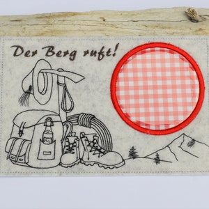 Peut inclure: Une carte en feutre blanc avec une broderie brune d'un sac à dos, de chaussures de randonnée, d'un chapeau et d'une corde. Le texte "Der Berg ruft!" est brodé en brun. Un cercle rouge avec un motif vichy blanc et rouge se trouve dans le coin supérieur droit.