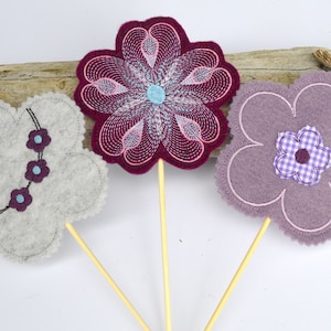 Pode incluir: Três enfeites de flores de feltro em palitos de madeira. As flores são em tons de roxo, cinza e rosa. A flor rosa tem um desenho de espiral branco e prateado. A flor roxa tem uma flor menor xadrez roxo e branco no centro. A flor cinza tem três pequenas flores roxas com centros azuis.