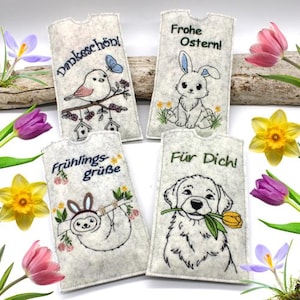 May include: Four felt pouches with embroidered designs. One features a bird and the text "Dankeschön!", another a bunny with "Frohe Ostern!", a sloth with "Frühlingsgrüße", and a dog with "Für Dich!". Spring flowers surround the pouches.