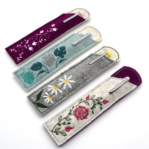 May include: Five felt pen holders with embroidered floral designs. The holders are in shades of purple, blue, gray, and white. Each holder has a pen inside.