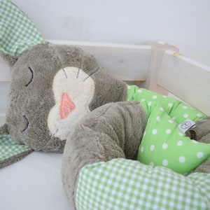 Puede incluir: Un juguete de peluche de conejo gris y verde con una cara blanca y una tela de lunares verde y blanco en la parte posterior. El conejo está acostado sobre una superficie blanca.
