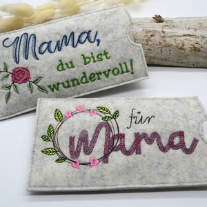 May include: Two gray felt pouches with embroidered designs. The first pouch has a rose and the text "Mama, du bist wundervoll!" The second pouch has a wreath with hearts and the text "f&uuml;r Mama".