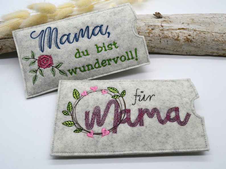 May include: Two gray felt pouches with embroidered designs. The first pouch has a rose and the text "Mama, du bist wundervoll!" The second pouch has a wreath with hearts and the text "f&uuml;r Mama".
