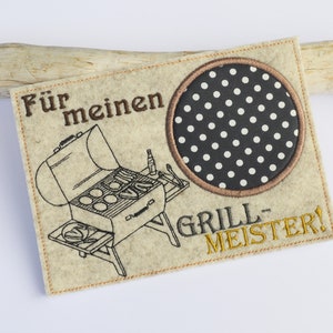 Peut inclure: Feutre beige brodé d'un cercle à pois noir et blanc, d'une illustration noir et blanc d'un barbecue avec de la nourriture et le texte "Für meinen Grill-Meister!"