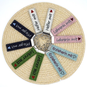 May include: A selection of felt keyrings in various colours, each with a silver-tone metal clasp. The keyrings feature embroidered text and small heart or heart-shaped designs. The keyrings are arranged on a woven, circular placemat.