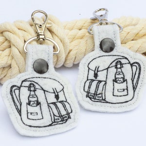 May include: Two white felt keyrings featuring a black outline design of a backpack with a bottle in the side pocket. Each keyring has a silver-coloured clasp and a dark grey snap closure. The keyrings are displayed on a cream-coloured rope.