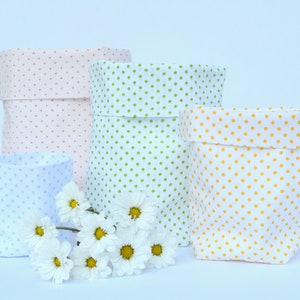 May include: Four fabric storage baskets with polka dot patterns in pink, green, blue, and yellow. The baskets are arranged in a row on a white surface with a few white daisies in front of them.