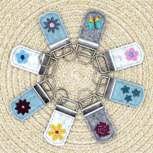May include: A collection of felt keychains with various embroidered floral and butterfly designs. The keychains feature a metal clasp and ring. The felt colors include blue, gray, and white, with flower designs in yellow, pink, and burgundy.