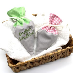 Puede incluir: Dos bolsas de tela gris con detalles verdes y rosas, atadas con cintas, descansan en una cesta de mimbre. Una tiene lunares verdes, la otra cuadros rosas. La primera bolsa tiene el texto bordado "Endlich Schulkind!"