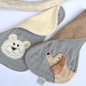 Könnte beinhalten: Zwei graue Baby-Spucktücher mit gestickten Tiergesichtern. Ein Tuch zeigt einen weißen Bären mit braunen Augen und einer schwarzen Nase. Das andere Tuch zeigt einen braunen Dackel mit braunen Augen und einer schwarzen Nase.
