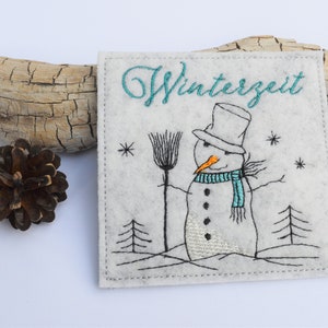 May include: Embroidered felt ornament featuring a snowman with a blue scarf and a broom, standing in a snowy landscape with trees. The text "Winterzeit" is embroidered in teal above the snowman.