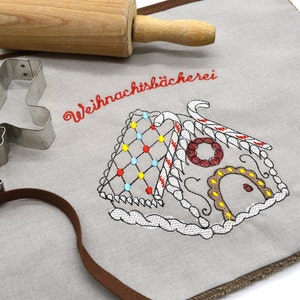 May include: A grey linen kitchen towel with a brown trim. The towel has an embroidered gingerbread house with red, yellow and blue candy decorations. The text "Weihnachtsbäckerei" is embroidered in red above the gingerbread house.