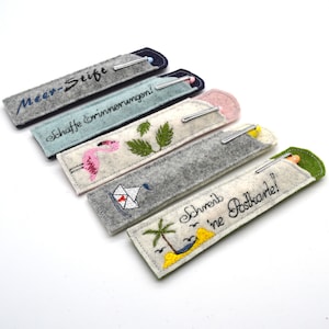 May include: Five felt pen holders with different embroidered designs. The designs include a flamingo, a sailboat, a palm tree, and the text "Meer-Stifte" and "Schreibe eine Postkarte!"