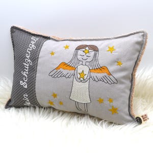 May include: A gray and white pillow with a brown trim. The pillow has an embroidered image of an angel with a star in its hands. The text "Dein Schutzengel" is embroidered on the side of the pillow.