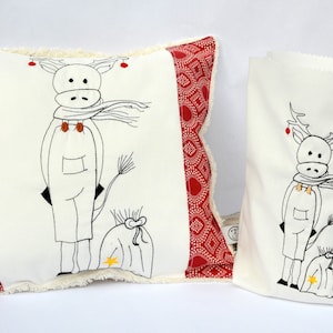 Può includere: Un cuscino bianco con un bordo a motivi rossi e bianchi presenta un disegno ricamato di una renna che indossa una sciarpa e porta un sacco. È mostrata anche una borsa bianca abbinata con un disegno ricamato simile.
