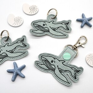 May include: Three blue whale keychains with black embroidered details. Each keychain has a silver ring and a silver clasp. The keychains are surrounded by seashells and starfish.