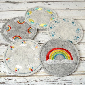 Peut inclure: Cinq dessous-de-verre ronds en feutre avec différents motifs brodés. Un dessous-de-verre présente un arc-en-ciel avec des nuages, un autre un arc-en-ciel avec des gouttes de pluie, un autre des étoiles, un autre des nuages et des gouttes de pluie et un autre un motif de paillettes dorées.