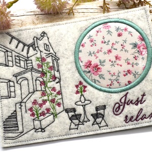 Peut inclure: Un dessous de verre en feutre gris clair avec un motif brodé. Le motif comprend un bâtiment avec des fleurs grimpantes, un cercle floral et les mots "Just relax!" en cursive. Le dessous de verre a une bordure cousue.