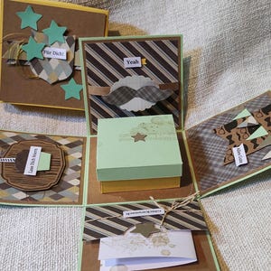 Könnte beinhalten: Eine handgefertigte Explosions-Geschenkbox mit einem zentralen hellgrünen und gelben Würfel, umgeben von vier Klappseiten, die mit braunen, gelben und grünen Mustern, Papiersternen, einem Schnurrbart-Ausschnitt und Etiketten mit deutschem Text verziert sind.