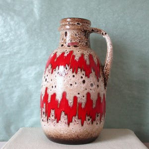Puede incluir: Un jarrón de cerámica vintage con asa, con una base crema con un patrón de zigzag rojo. El jarrón tiene un borde y un asa marrones, y está salpicado de manchas oscuras. Una pieza decorativa para la decoración del hogar.