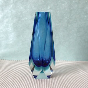 Peut inclure: Un vase en verre haut et effilé présentant un centre bleu profond qui se transforme en verre transparent et angulaire à la base. Le design comprend des indentations verticales incurvées qui créent une sensation de profondeur et de mouvement.