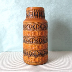 Può includere: Un vaso in ceramica arancione di forma cilindrica. Il vaso presenta un motivo ripetuto di forme geometriche marrone scuro, tra cui cerchi, triangoli e rettangoli. Il vaso ha una superficie strutturata e una parte superiore leggermente svasata.