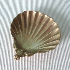 Può includere: Un piatto in metallo color oro a forma di conchiglia di capasanta. L'interno presenta profonde scanalature radianti che si estendono dalla base fino al bordo smerlato. La base è decorata con un piccolo dettaglio metallico con un elemento circolare.