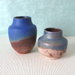 Può includere: Due vasi in ceramica con una smaltatura blu e marrone. Il vaso più grande è di un blu più scuro con un fondo marrone, mentre il vaso più piccolo è di un blu più chiaro con un fondo marrone e bianco.
