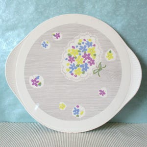 Könnte beinhalten: Ein weißes Serviertablett mit floralem Design. Das Tablett zeigt einen Strauß gelber, violetter und blauer Blumen auf grauem Hintergrund. Zusätzliche verstreute Blumen in den gleichen Farben sind ebenfalls vorhanden. Das Tablett hat einen weißen Rand und Griffe.