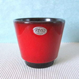 Op de afbeelding: Een rode keramische pot met een zwarte rand en een wit label met de tekst "Jopcko West-Germany Keramik".