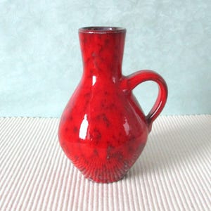 Könnte beinhalten: Eine kleine rote Keramikvase mit Henkel. Die Vase hat eine rot gefleckte Glasur und einen leicht ausgestellten Rand.