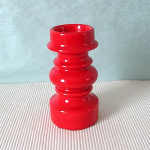 May include: A bright red candle holder with a glossy finish. The ceramic piece features a rounded base, a series of stacked, bulbous sections, and a flared top. The design is symmetrical and eye-catching. A decorative item for the home.