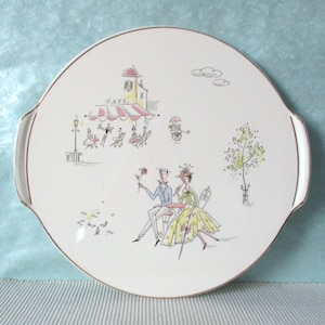 Könnte beinhalten: Eine Vintage-Servierplatte mit einer skurrilen Illustration einer Pariser Cafészene. Die cremefarbene Platte zeigt ein Café mit einer rosa-weiß gestreiften Markise, Figuren und dem Wort "CAFE". Der Rand ist mit Gold verziert.