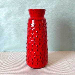 Può includere: Un vaso alto in ceramica rossa con una superficie strutturata da piccole rientranze scure. Il vaso ha una parte superiore svasata e un corpo cilindrico. Questo vaso decorativo è un pezzo d'arredo di grande effetto.
