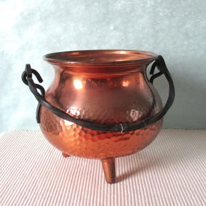 Könnte beinhalten: Ein Kupferkessel mit gehämmerten Texturen und drei kleinen Beinen. Der Kessel hat einen schwarzen, gebogenen Griff und einen abgerundeten Körper. Das Kupfer hat eine glänzende, reflektierende Oberfläche.