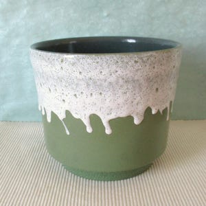 Può includere: Un vaso di ceramica a due toni. La metà inferiore è verde pieno, mentre la metà superiore presenta una glassa bianca maculata che cola sui lati. L'interno è verde scuro.