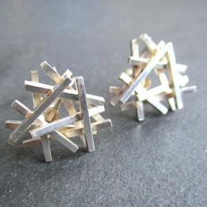 May include: A pair of silver stud earrings. Each earring features a cluster of rectangular bars. The earrings have a geometric, modern design and are displayed on a dark grey surface. The earrings are made of silver.